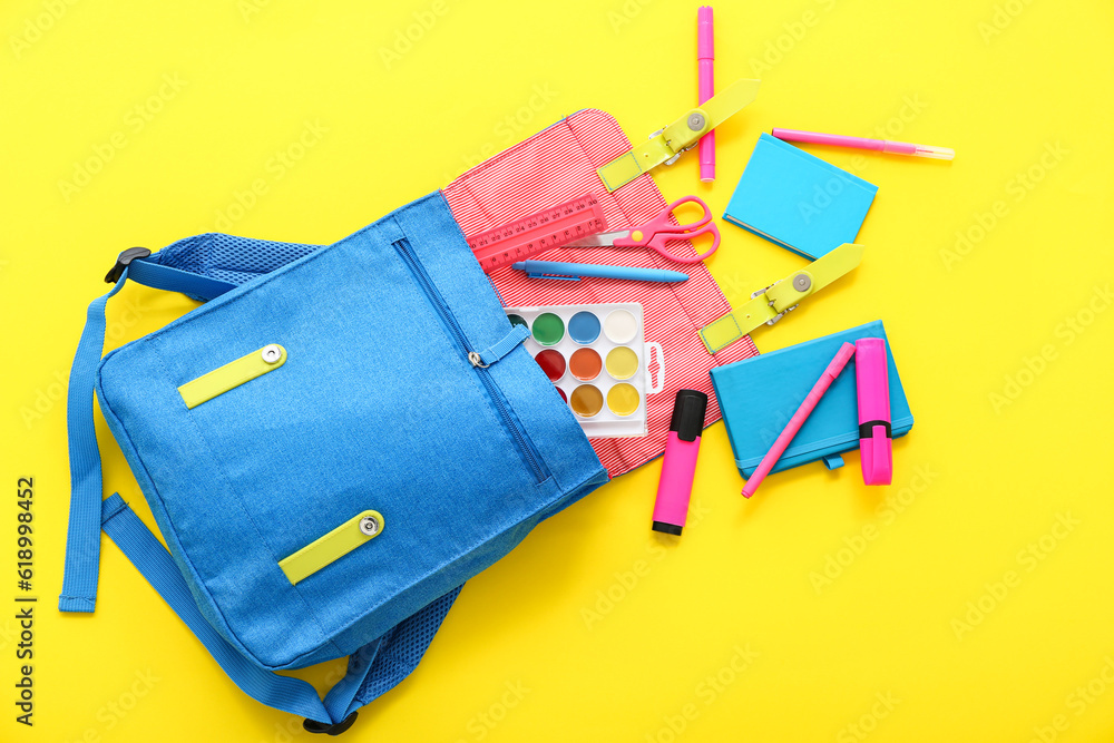 Blue school backpack with notebooks, markers and watercolors on yellow background