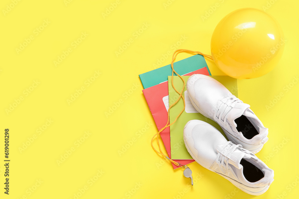 Sneakers with whistle, ball and notebooks on yellow background