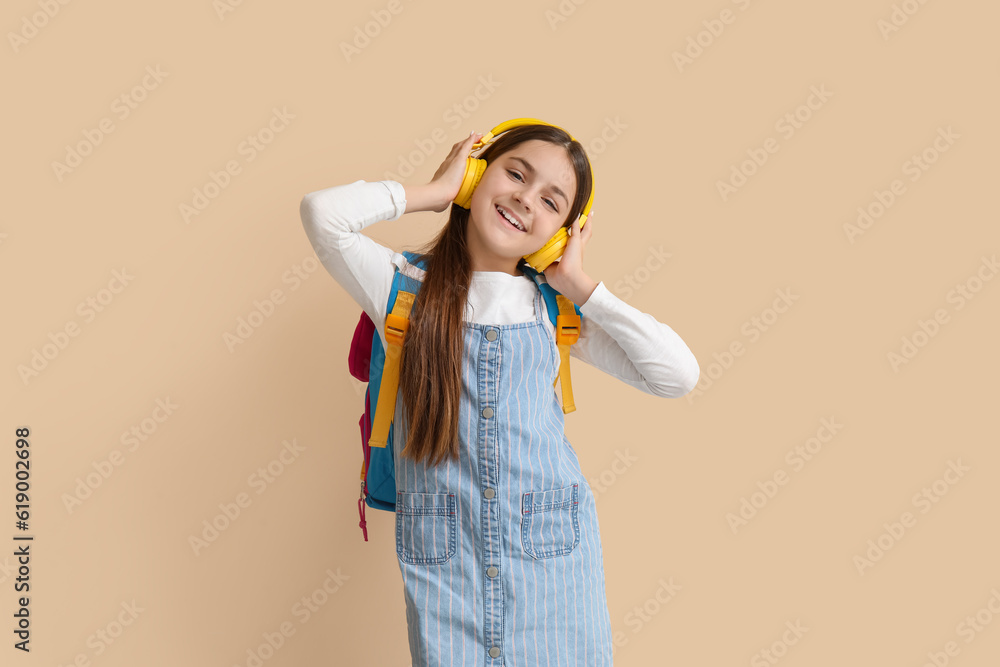 Little schoolgirl in headphones with backpack on beige background