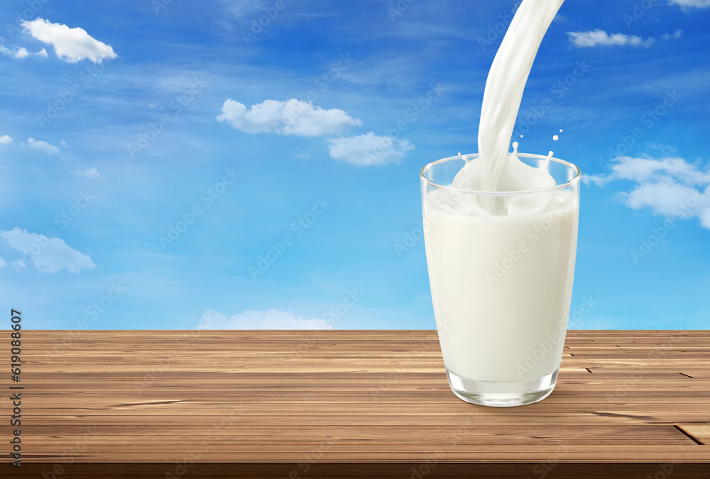 Pouring milk into glass on wooden table with bright sky as background