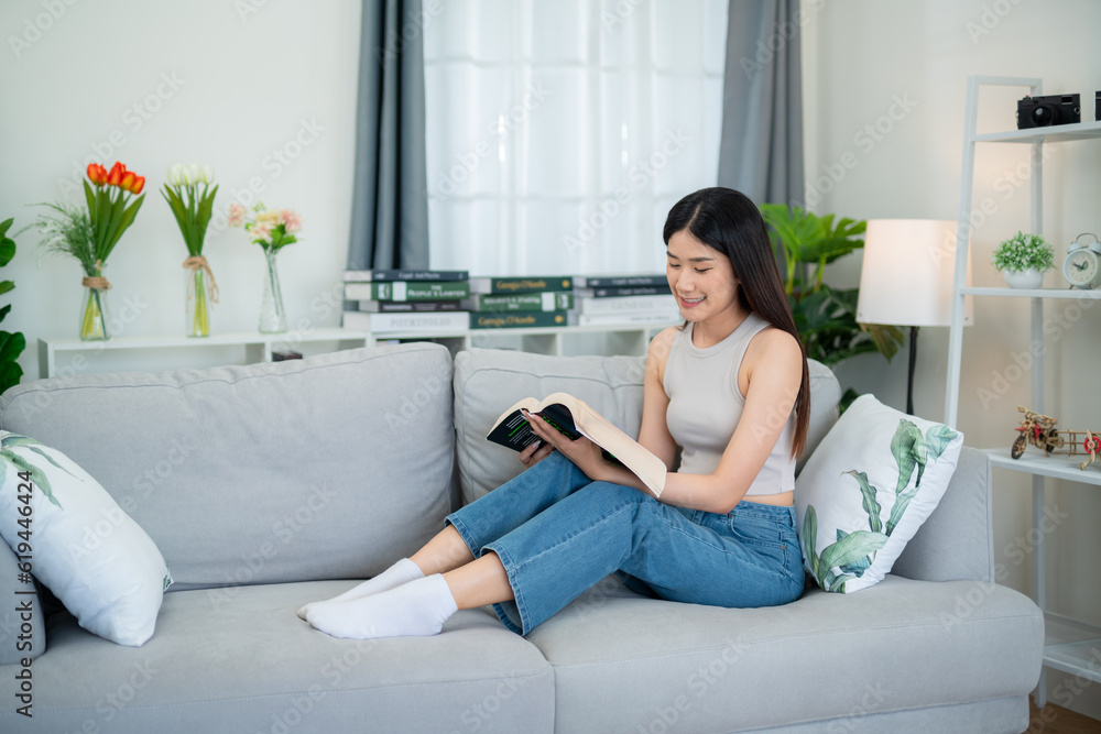 Asian young woman sitting on the sofa in the living room reading a book happily on weekends.