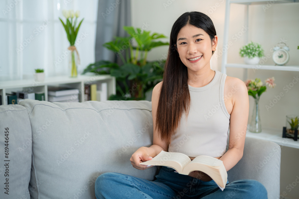 Smiling asian young woman looking at the camera while happily reading a book on the sofa in the livi