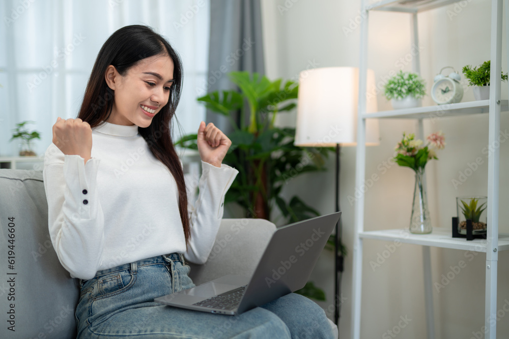 Asian woman showing success gesture while using laptop on sofa in living room.