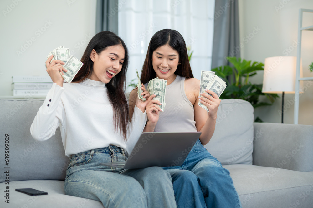 Two young asian women holding cash and successful online shopping with laptop in living room. Financ