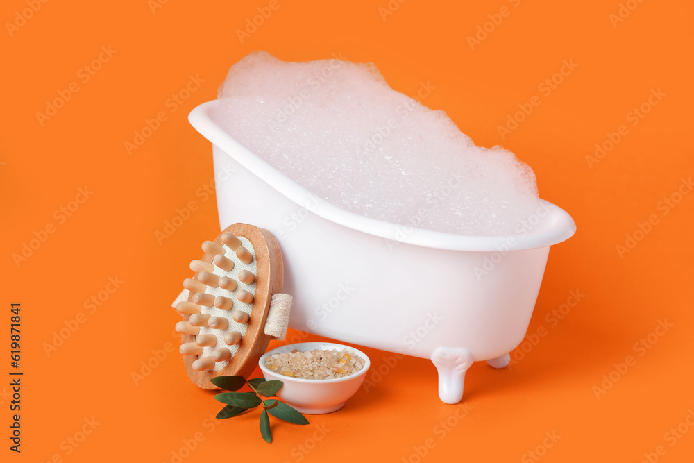 Small bathtub with foam, massage brush and sea salt on color background