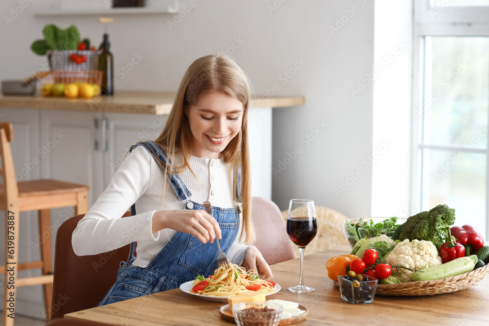 Young woman with glass of wine eating tasty pasta in kitchen