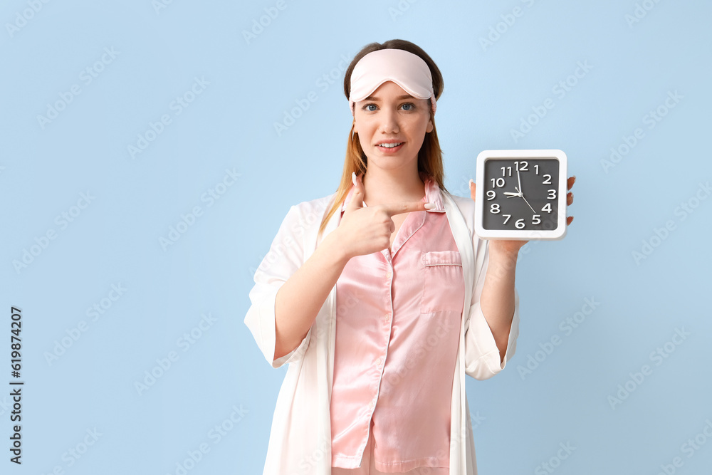 Young woman in pajamas pointing at clock on blue background