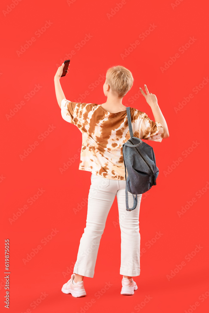 Female student with backpack taking selfie on red background, back view