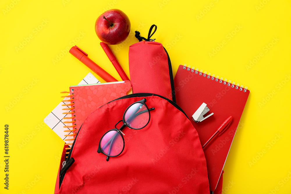 Red school backpack with notebooks, eyeglasses and pencil case on yellow background