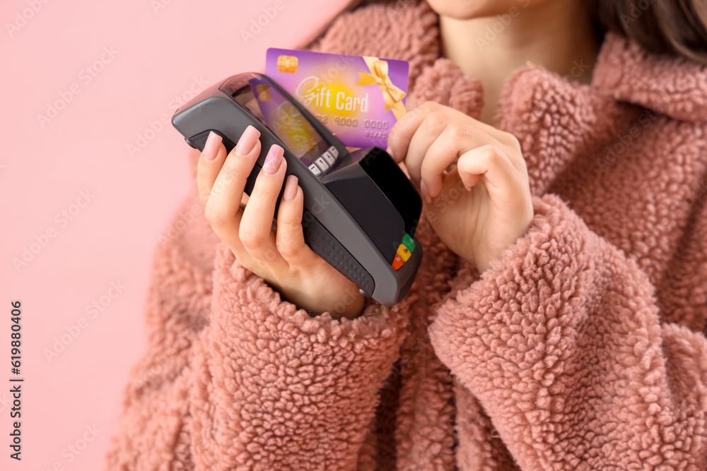 Young woman with payment terminal and gift card on pink background, closeup