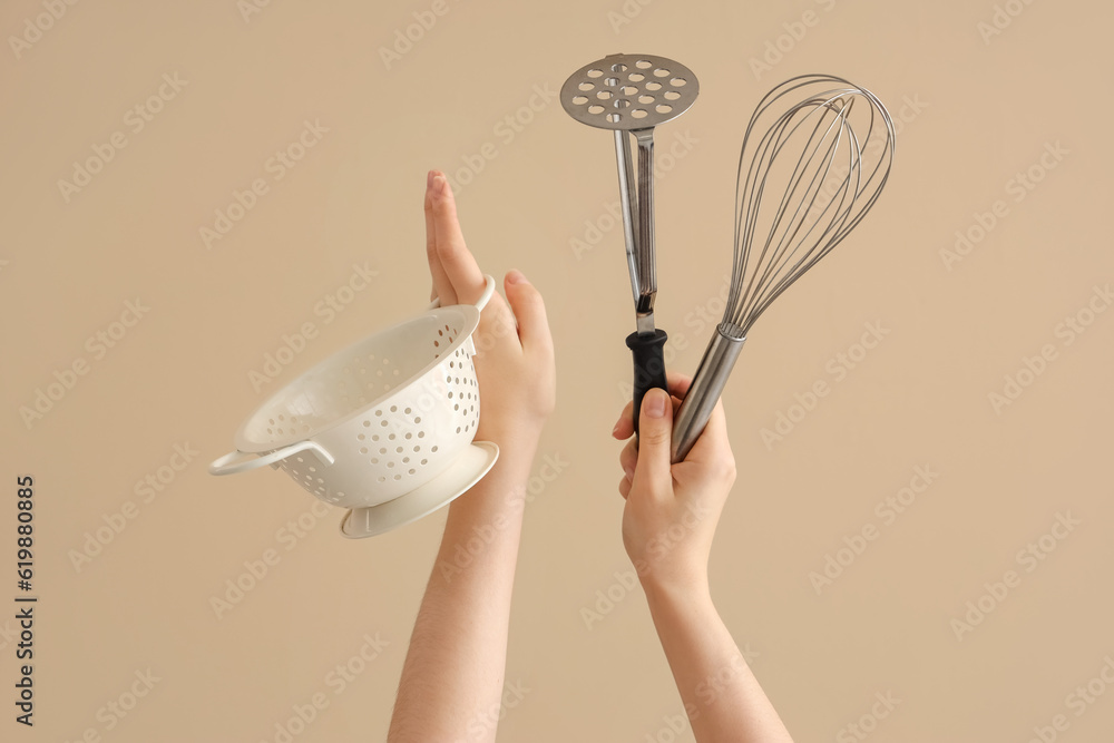 Female hands holding colander, whisk and potato masher on brown background