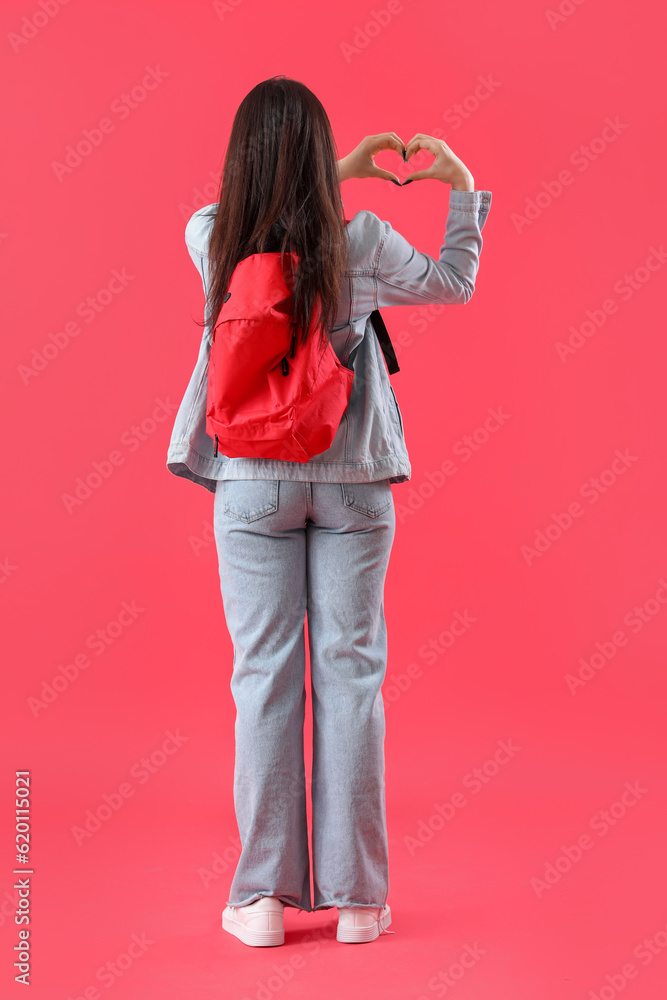 Female student with backpack making heart shape on red background, back view