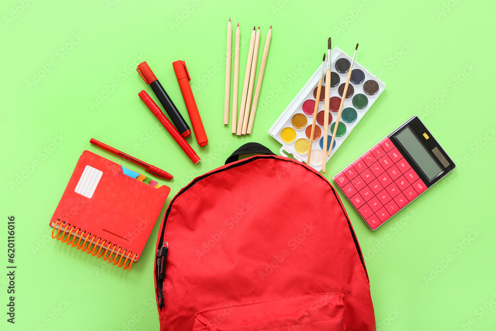 Red school backpack with calculator, color pencils and watercolors on green background