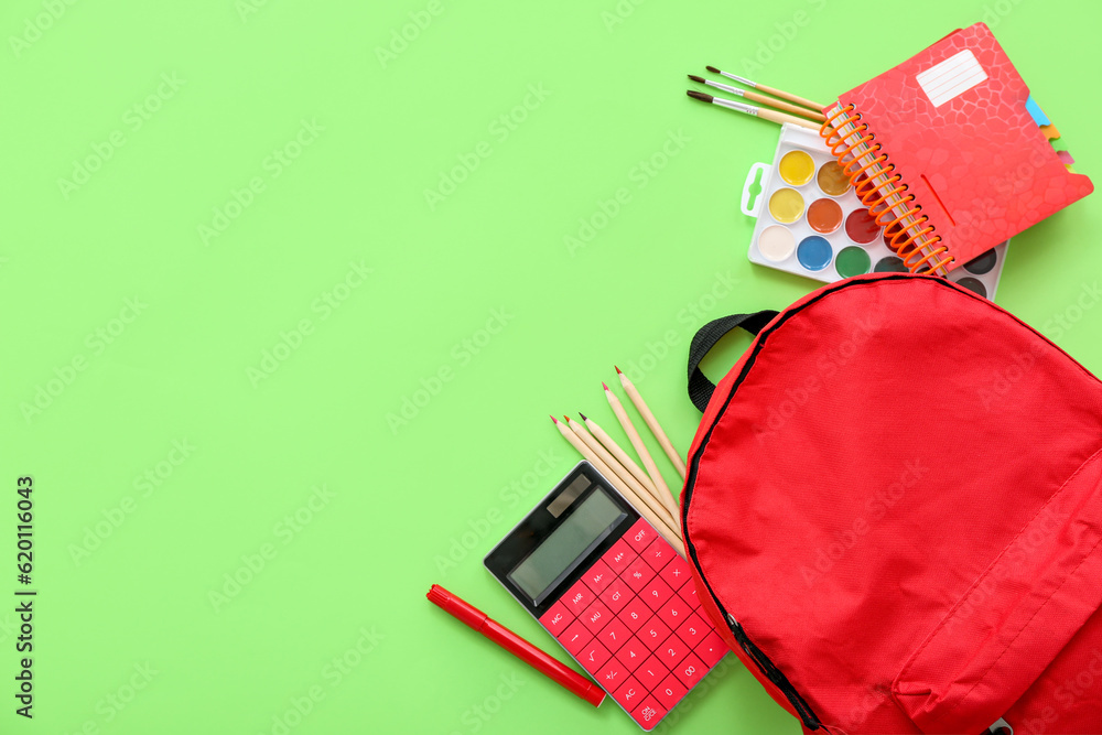 Red school backpack with calculator, color pencils and watercolors on green background