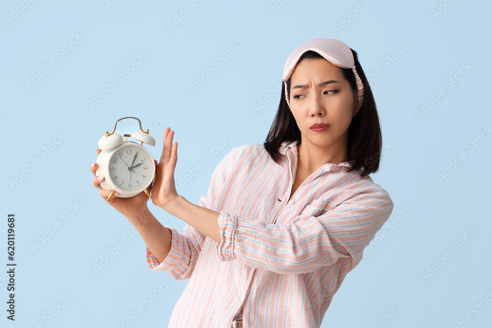 Displeased Asian woman in pajamas with alarm clock on blue background