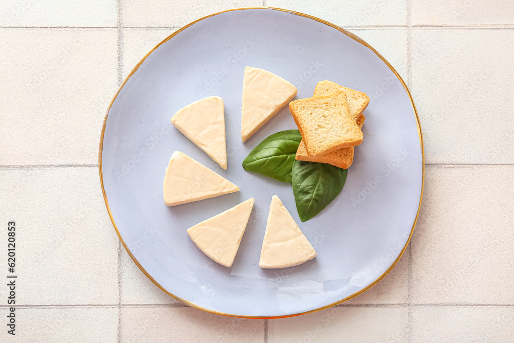 Plate with triangles of tasty processed cheese, basil leaves and croutons on light tile background