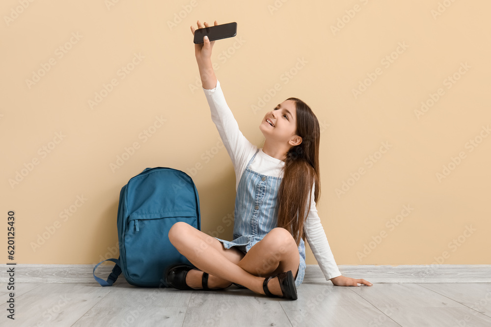 Little schoolgirl with backpack taking selfie near beige wall