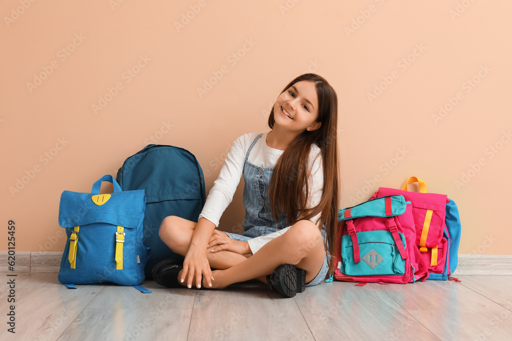 Little schoolgirl with backpacks sitting near beige wall