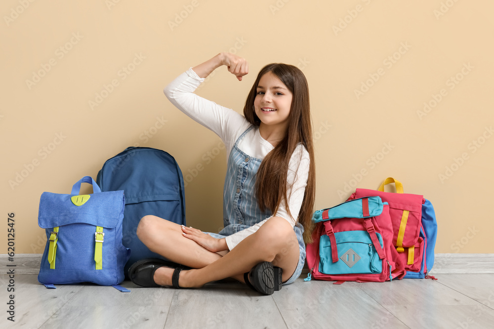 Little schoolgirl with backpacks showing muscles near beige wall