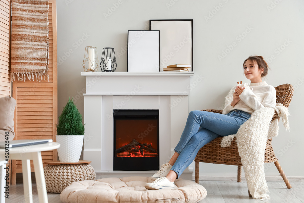 Young woman with cup of hot cocoa sitting in armchair near fireplace at home