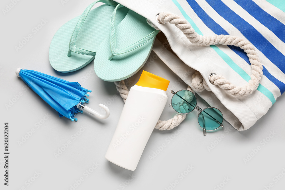 Bag with sunglasses, flip flops, mini umbrella and bottle of sunscreen cream on grey background