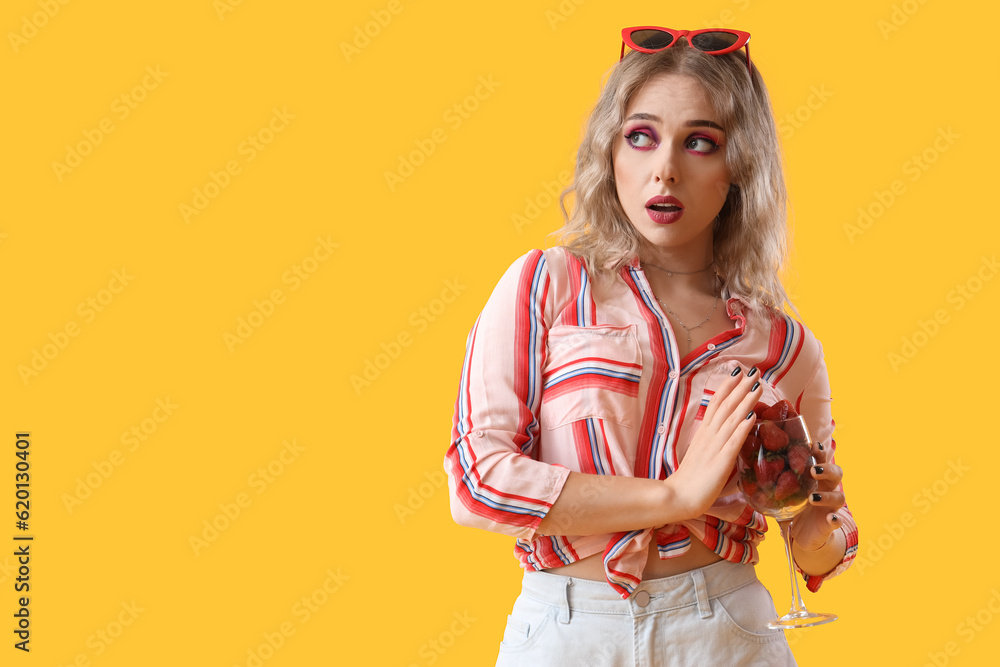 Upset young woman with glass of strawberries on yellow background