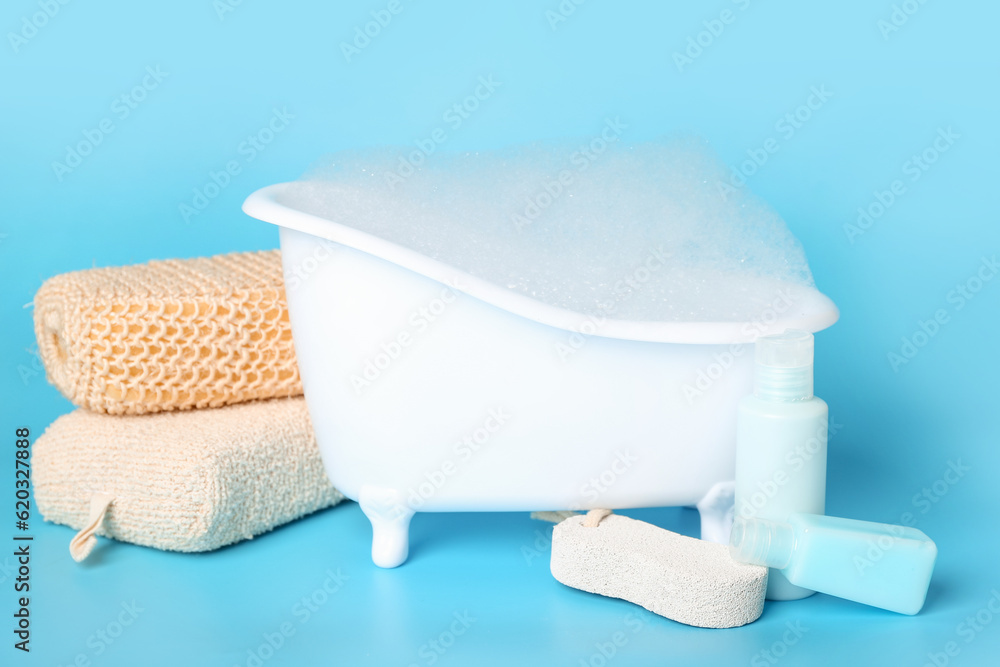 Small bathtub with foam and bath supplies on color background