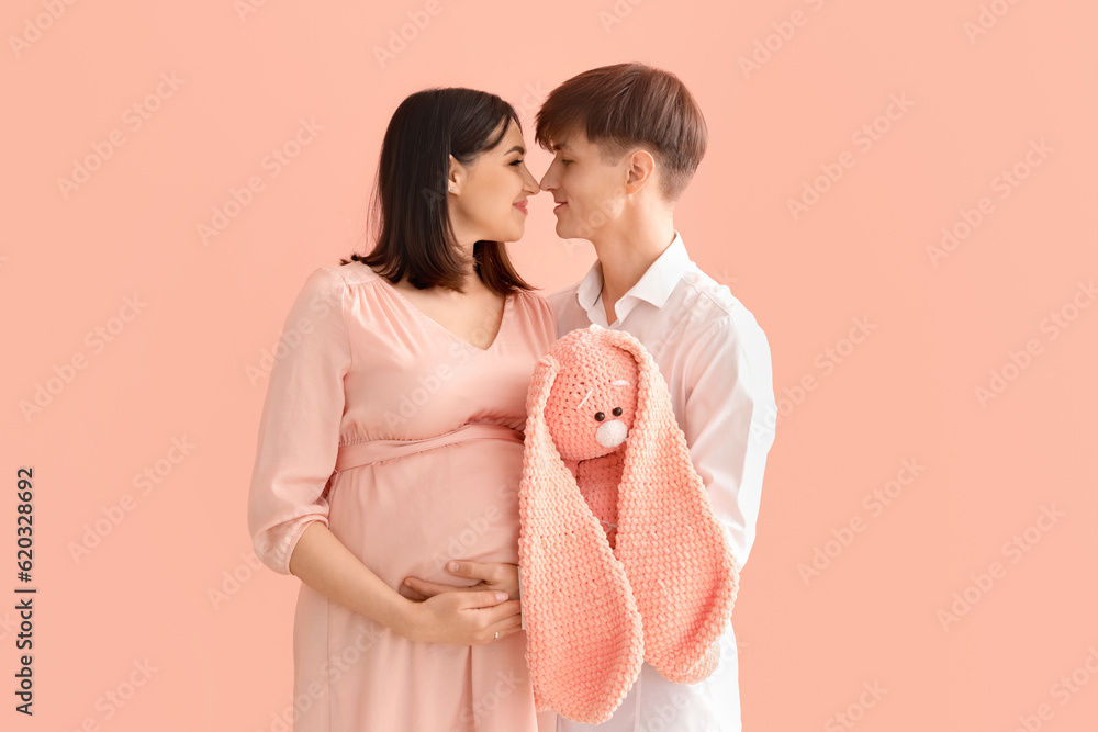 Young pregnant couple with toy bunny on pink background