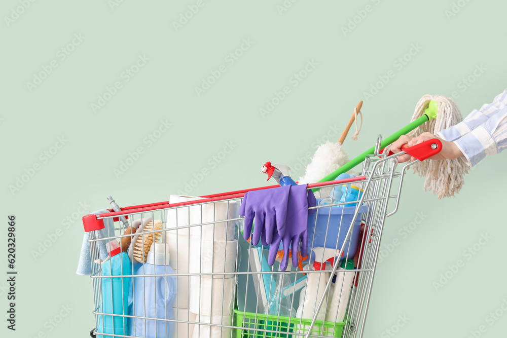 Woman with shopping cart full of cleaning supplies on pale green background