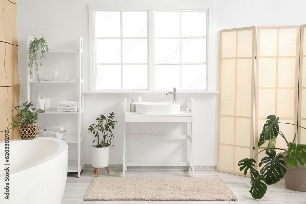 Interior of light bathroom with sink, bathtub, shelving unit and houseplants