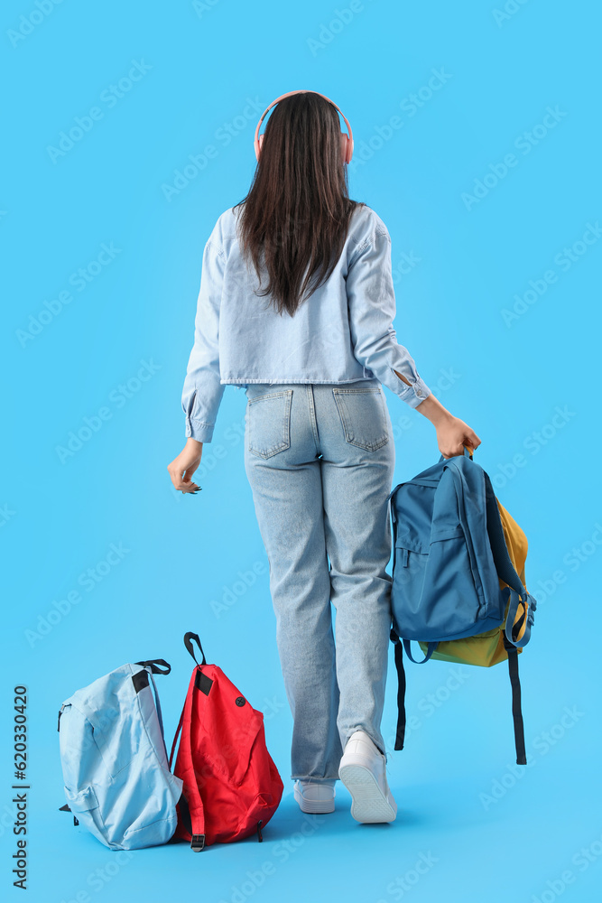 Female student in headphones with backpacks on blue background, back view