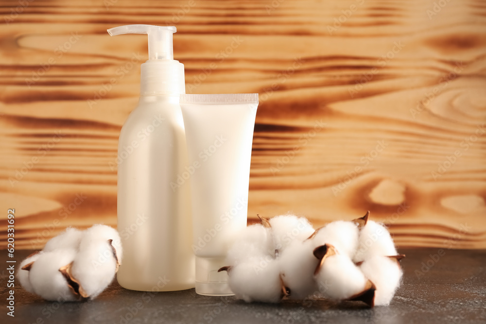 Bottle of cosmetic products and cotton flowers on dark table against wooden background