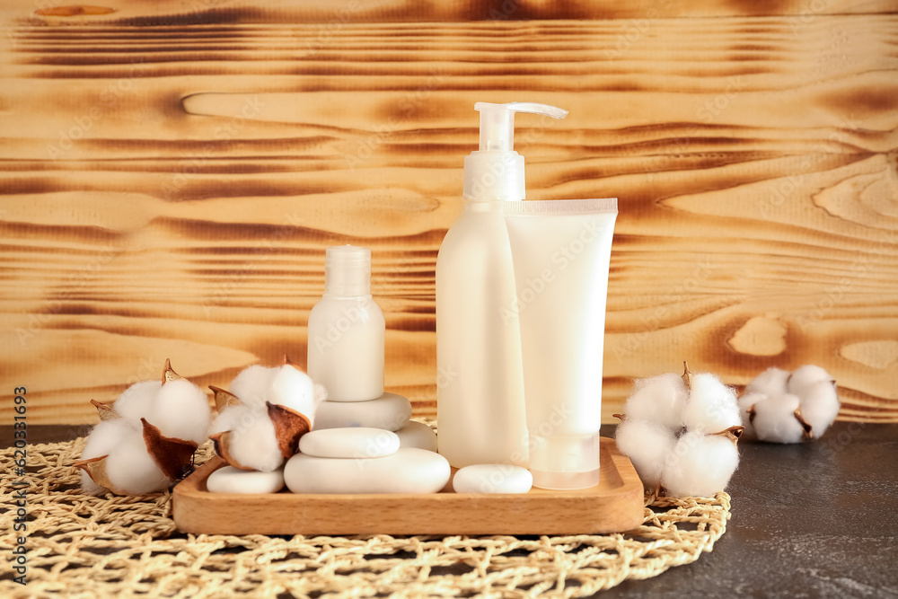 Bottles of cosmetic products, spa stones and cotton flowers on table against wooden background