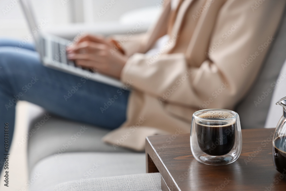 Woman sitting on sofa and using laptop near table with glass of delicious coffee