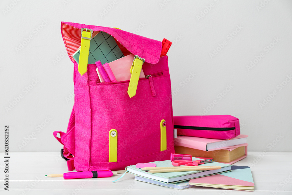 Pink school backpack with books, pencil case and stationery on white wooden table near wall