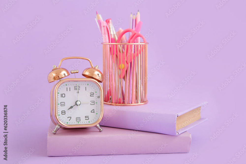 Alarm clock with books and stationery holder on lilac background