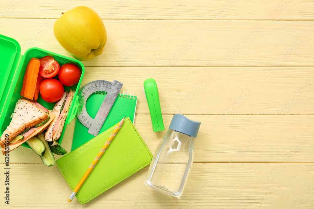 Stationery, drink and lunch box with tasty food on yellow wooden background