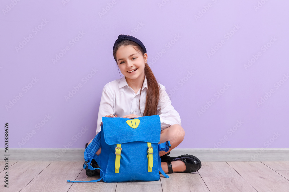 Little schoolgirl with backpack sitting near lilac wall