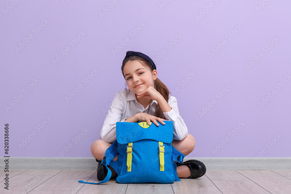 Little schoolgirl with backpack sitting near lilac wall