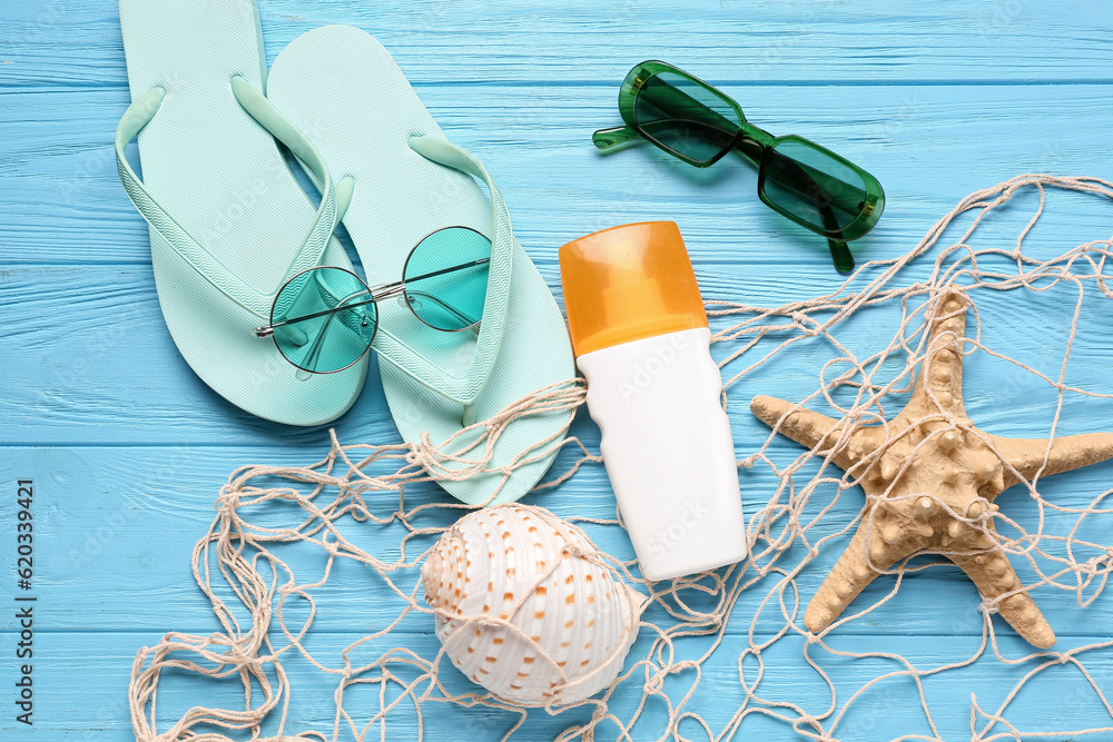 Net with flip flops, sunglasses and bottle of sunscreen cream on blue wooden background