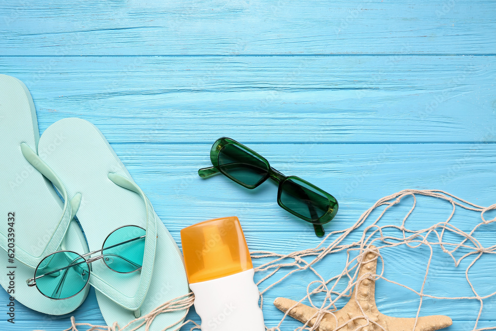 Net with flip flops, sunglasses and bottle of sunscreen cream on blue wooden background