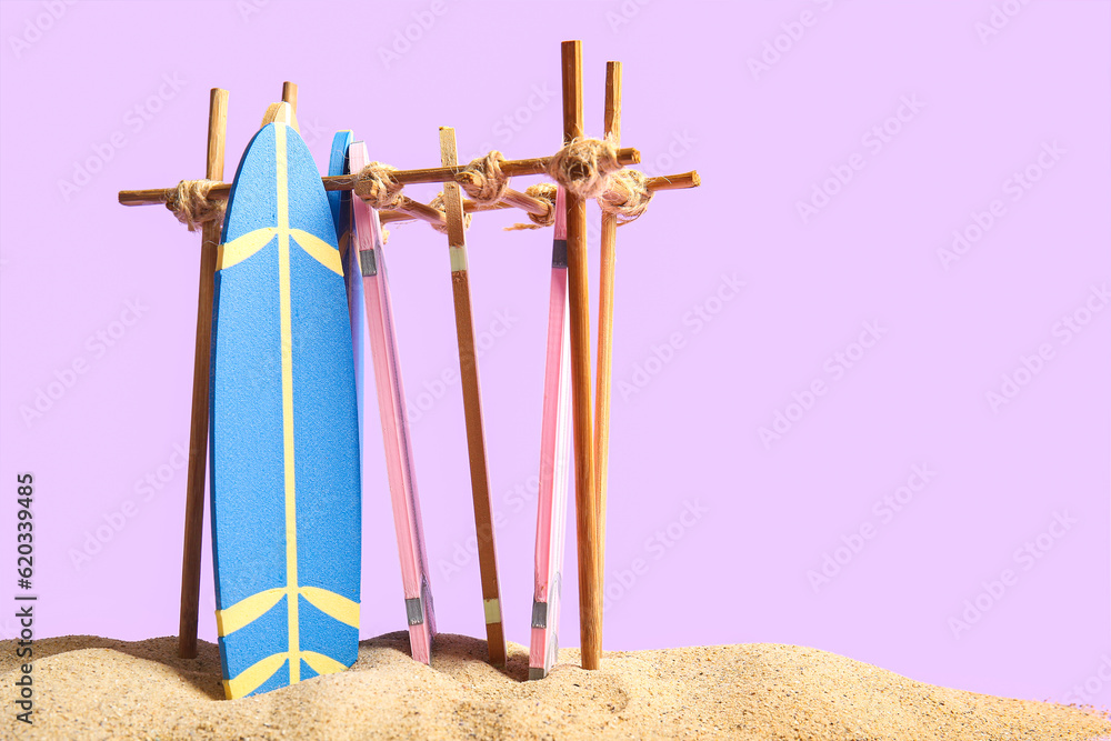 Stand with different mini surfboards on sand against lilac background