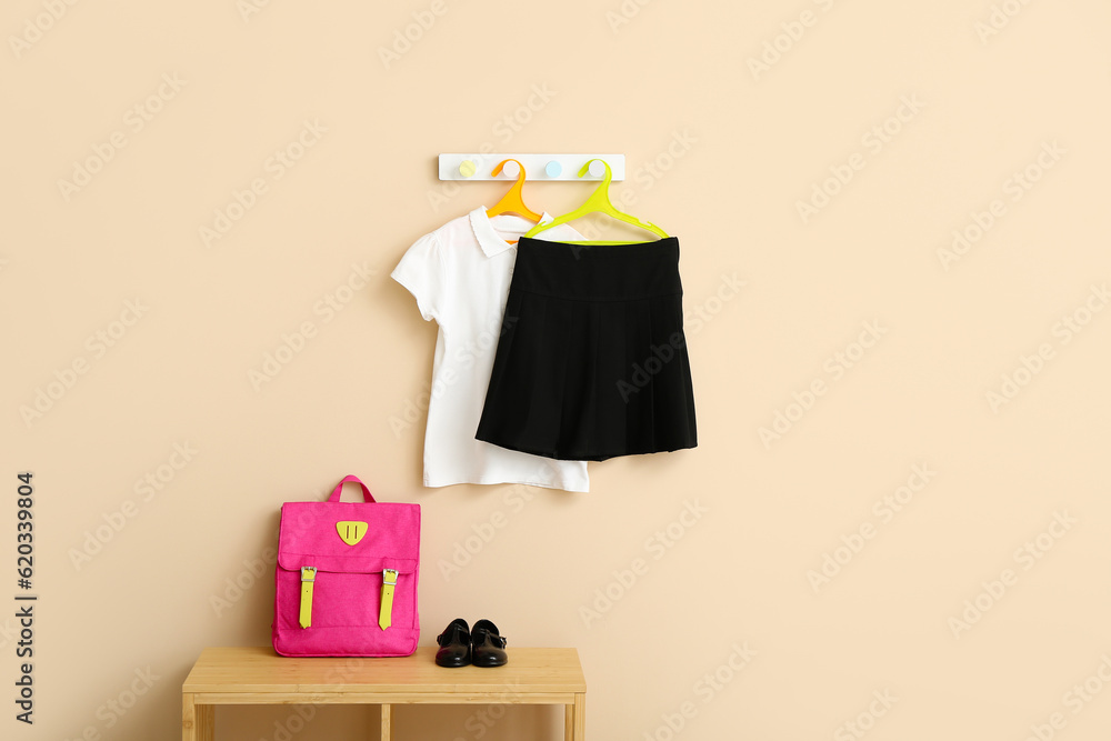 Stylish school uniform hanging on rack with backpack against beige wall