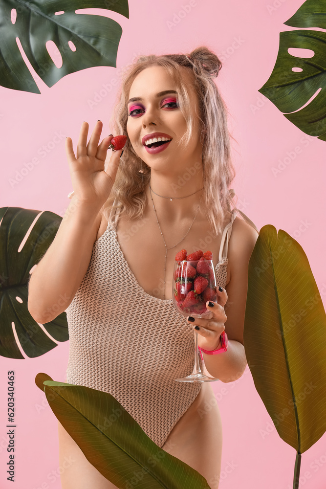 Young woman in swimsuit with glass of strawberries and palm leaves on pink background