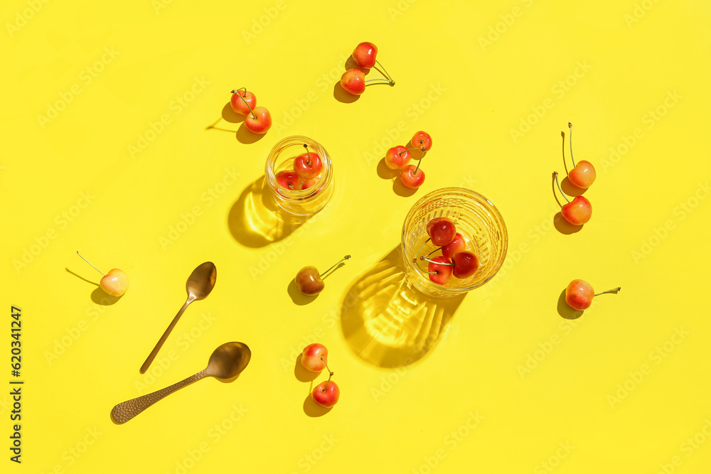 Glass and jar with sweet cherries on yellow background