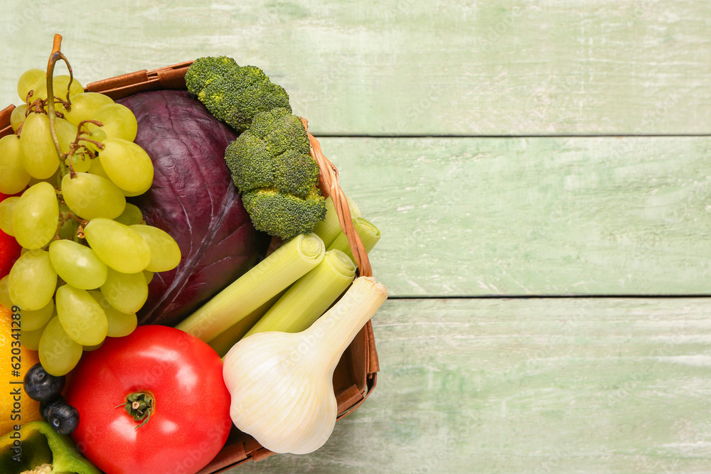 Wicker basket with different fresh fruits and vegetables on green wooden background, closeup