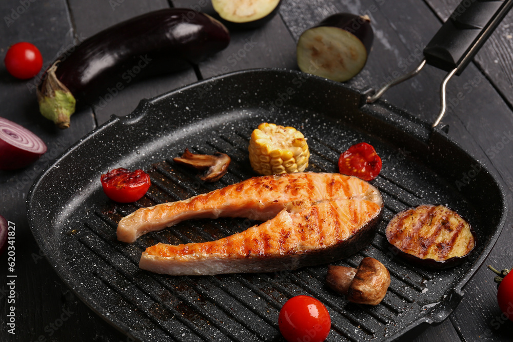 Frying pan with tasty grilled salmon steak and vegetables on black wooden background