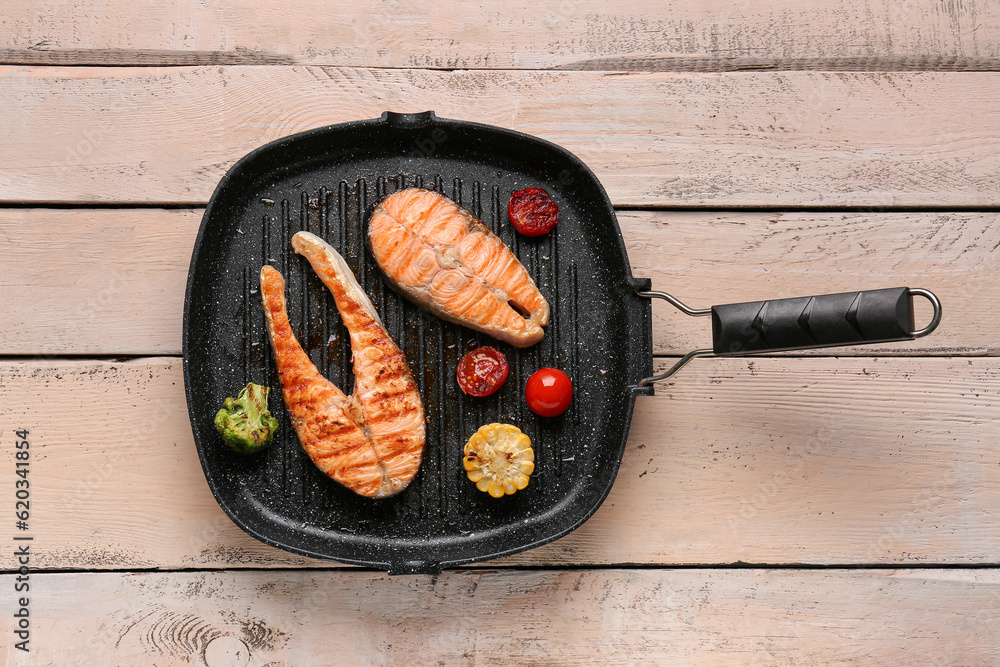 Frying pan with tasty grilled salmon steaks and vegetables on light wooden background