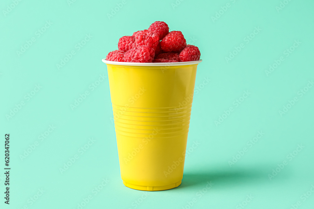 Plastic cup with fresh raspberries on turquoise background