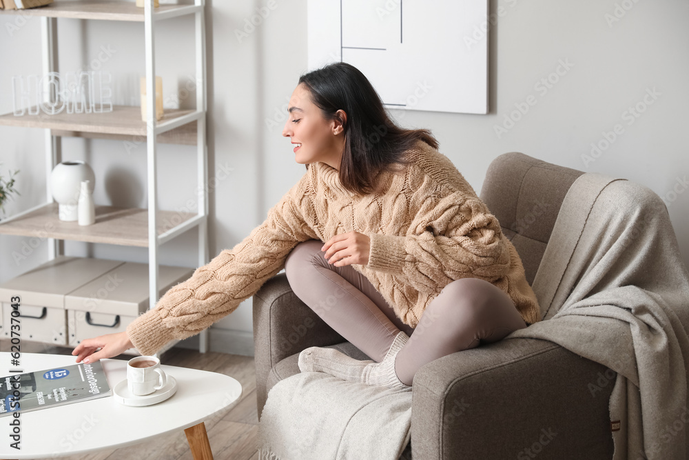 Young woman in soft armchair taking magazine from table at home